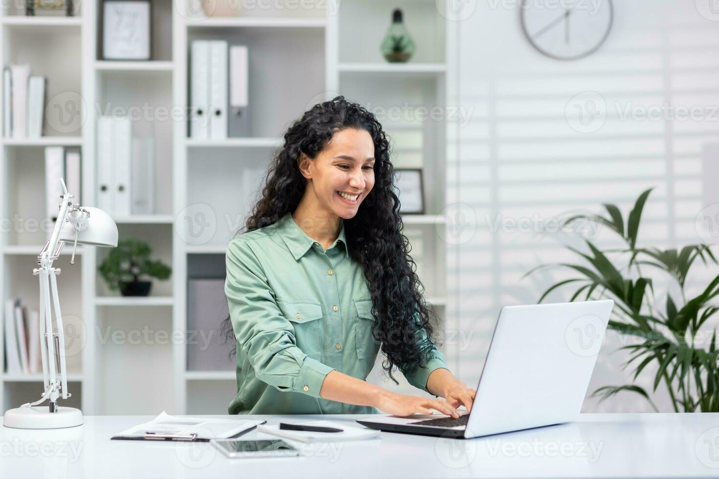 Young successful hispanic businesswoman working in bright home office indoors sitting at table with documents and reports, woman using laptop at work happy smiling. photo