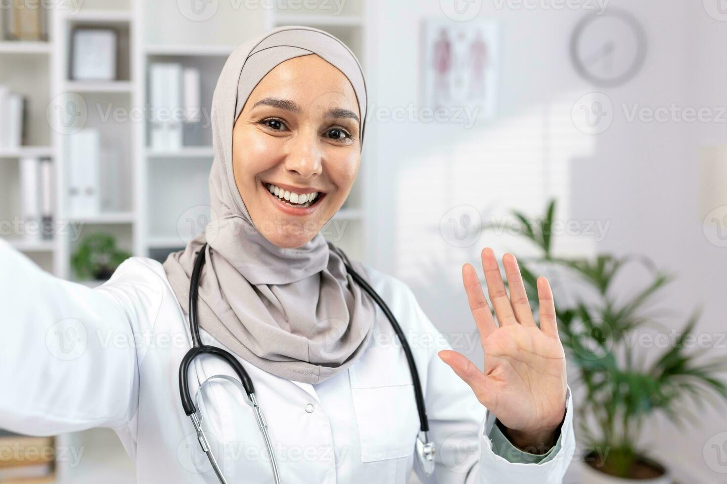 Beautiful asian muslim female doctor in hijab and suit taking selfie photo while resting. A doctor makes a video call, smiling and waving as he greets his family from his workplace