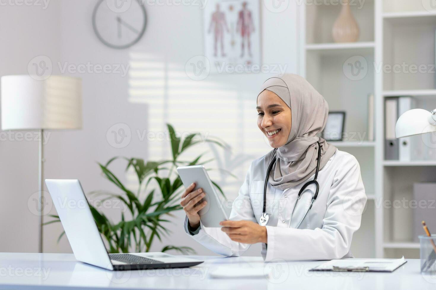 Portrait Muslim female doctor in hijab, woman doctor with tablet computer using application, smiling satisfied work working inside medical office, in white medical coat, sitting at table with laptop photo
