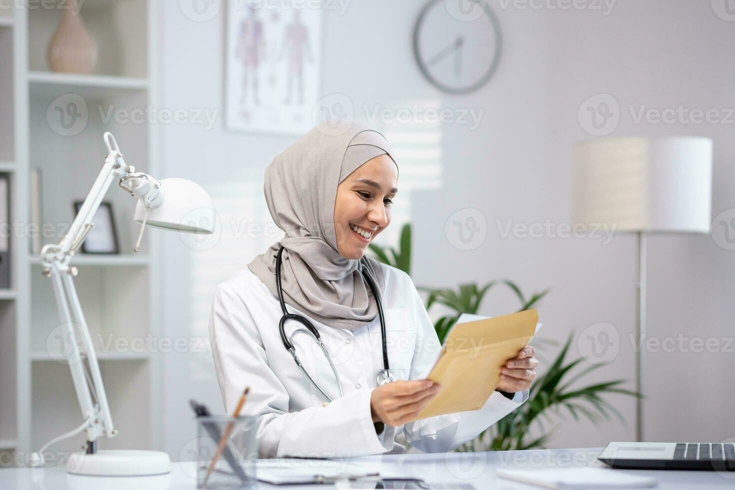 Joyful female doctor in hijab received envelope message mail notification with good results of patient's treatment, woman is happy and smiling, working inside clinic office in white medical coat. photo