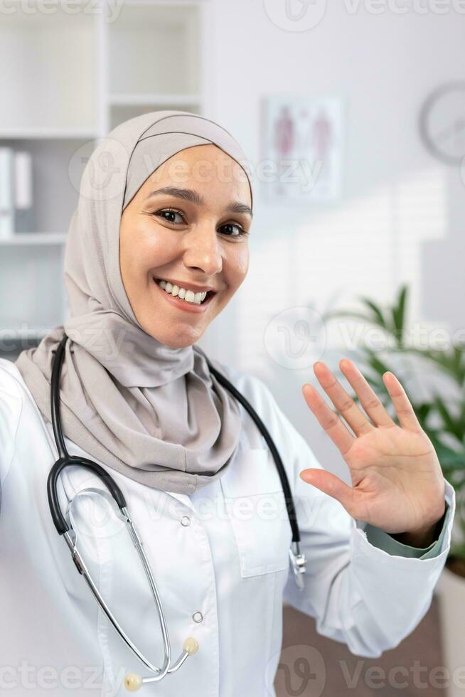 A Muslim female doctor in a hijab conducts an online appointment with patients on the phone, waves to the camera, view from the phone. photo