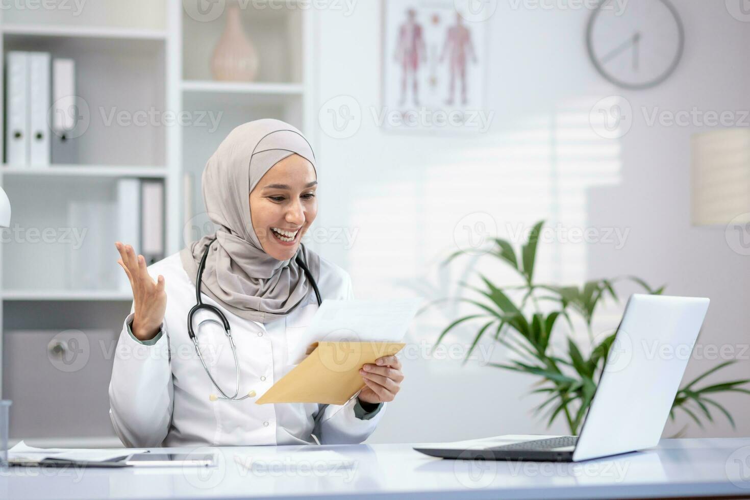 Happy female doctor in hijab holding letter in hands, sitting at table in hospital office, happy with good news. photo