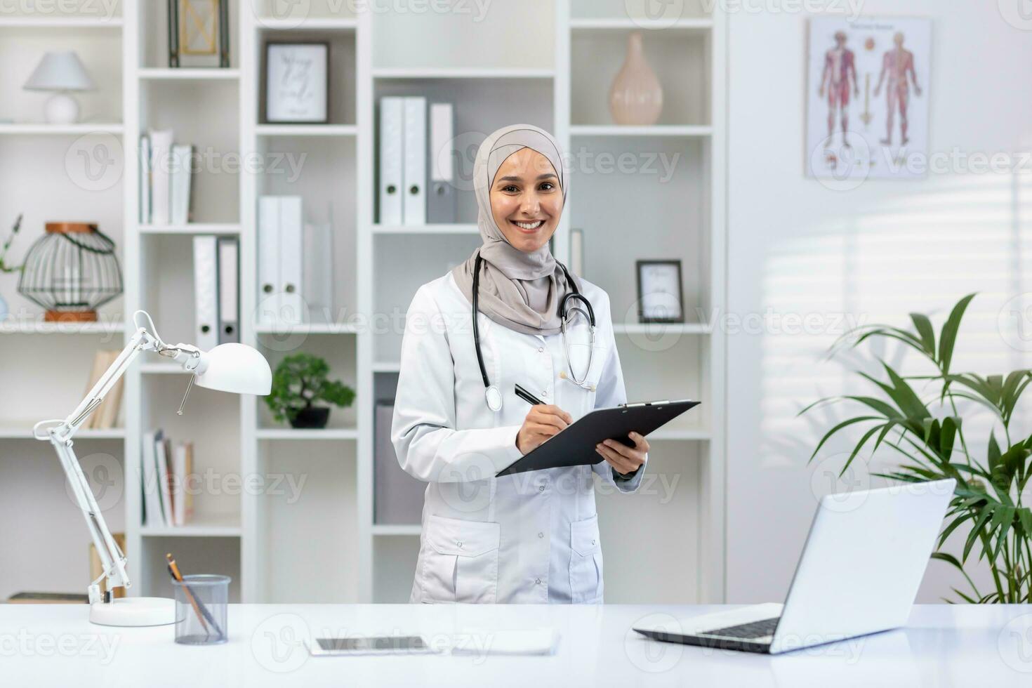 Portrait of a Muslim female doctor in a hijab standing in a modern office, holding a tablet with documents in her hands, smiling and looking at the camera. photo