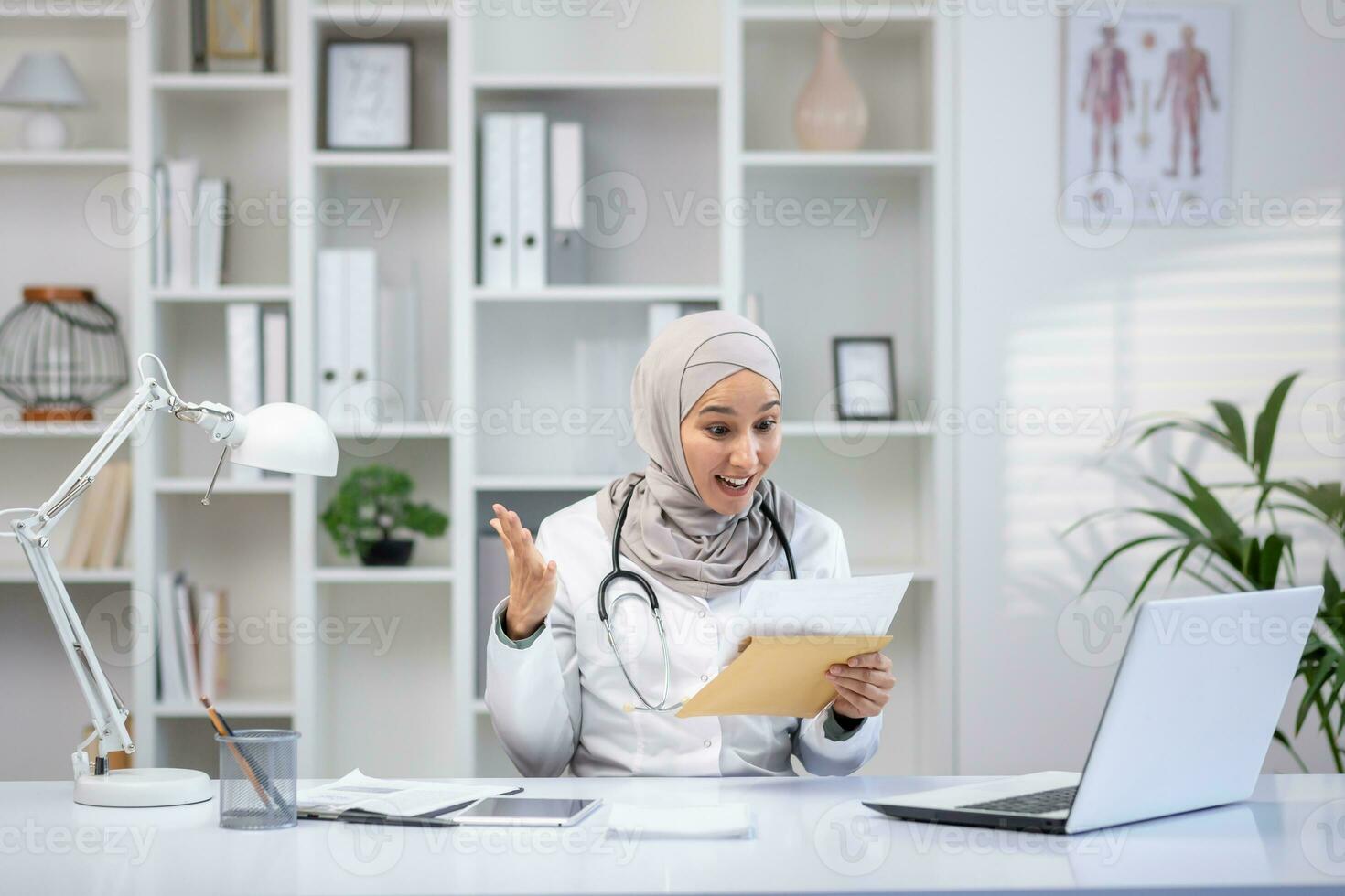 Joyful female doctor in hijab received envelope message mail notification with good results of patient's treatment, woman is happy and smiling, working inside clinic office in white medical coat. photo