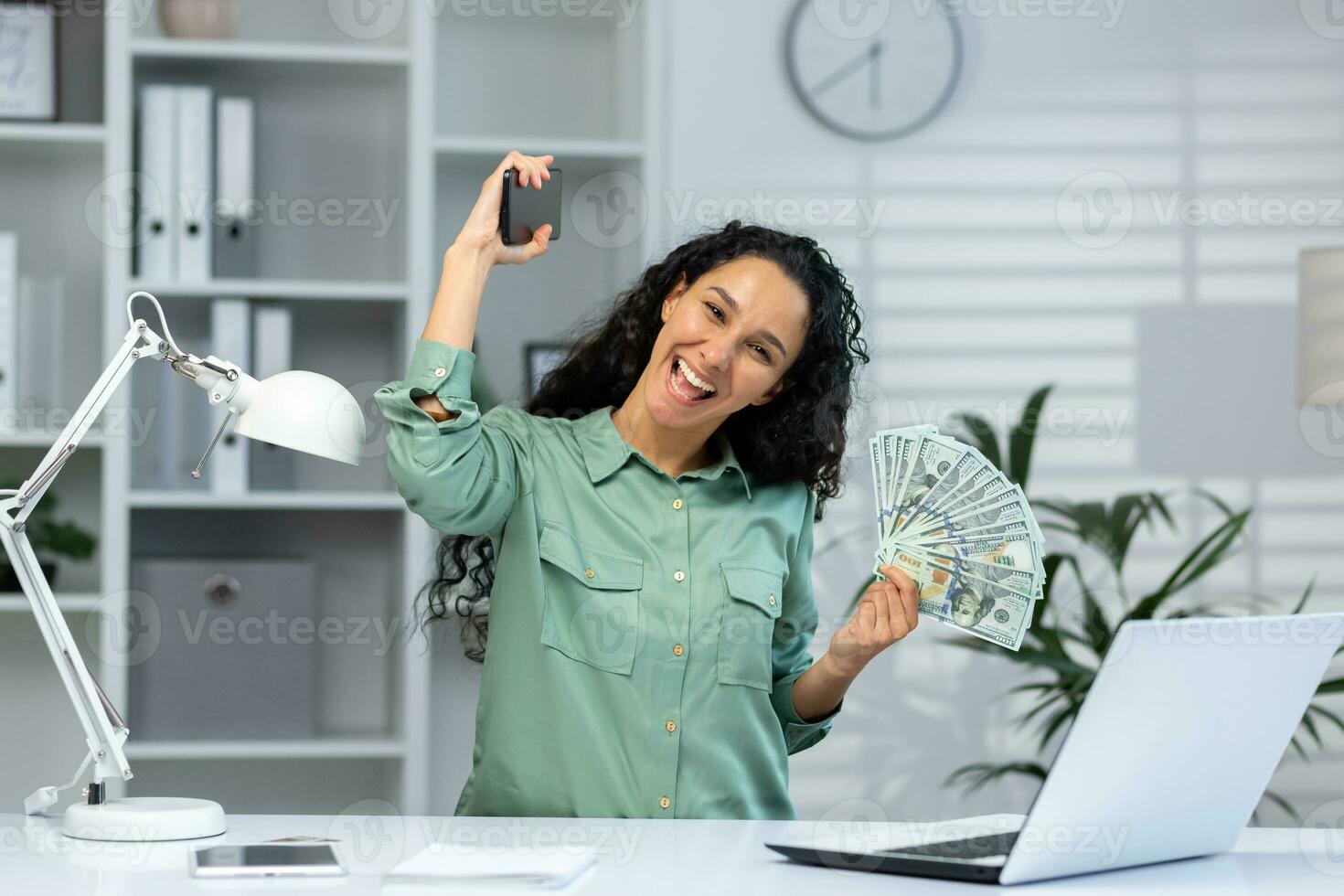 retrato de alegre contento y exitoso negocio mujer dentro oficina, Hispano mujer sonriente participación teléfono y americano dolares dinero efectivo ganancia. foto