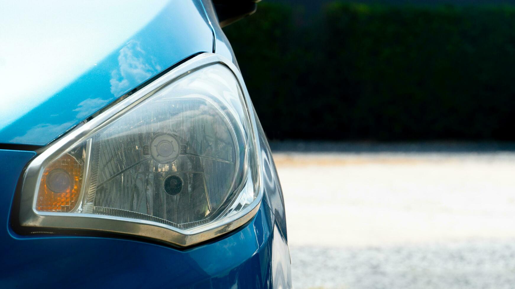 Headlights of bue car. with sunlight casting a radiant glow on the hood. The background depicts a blurred parking lot and dark green trees. photo