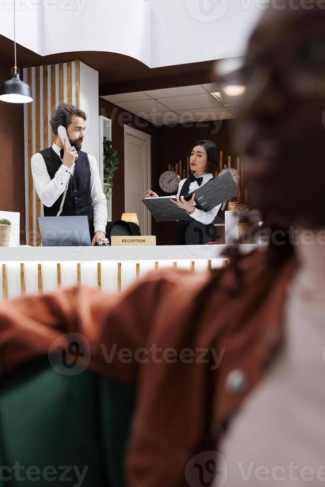 Hotel staff working on reservations at front desk with records, using landline phone to make online bookings at reception counter in lobby. Man and woman receptionists doing check in. photo