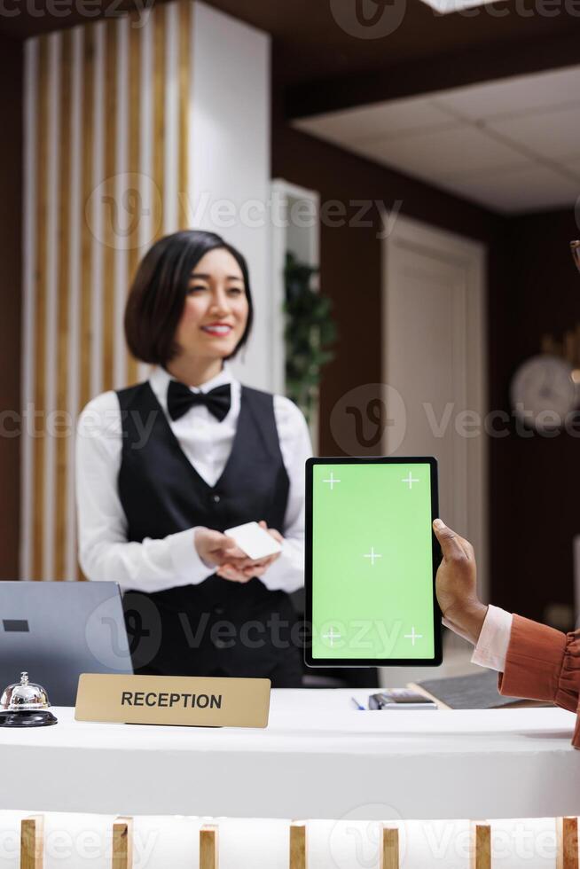 mujer participación tableta con pantalla verde a frente escritorio, recepción habitación acceso tarjeta desde asiático recepcionista. joven persona utilizando aislado Bosquejo monitor con chromakey a hotel recepción. foto