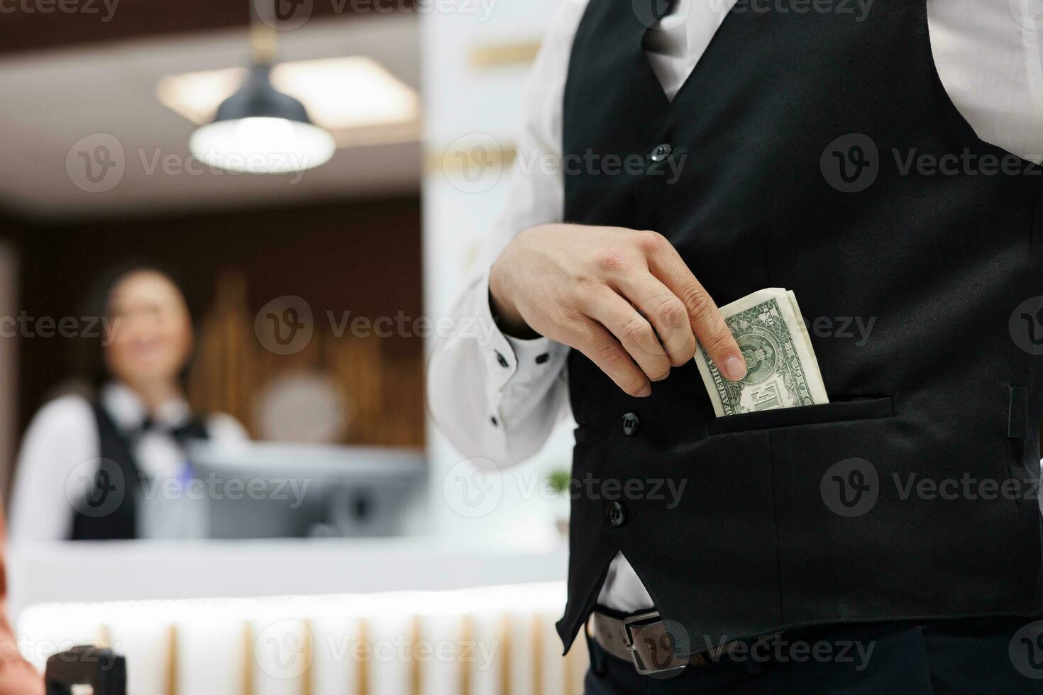 Hotel staff receives money in lobby, putting cash in pocket to help guests with luggage after check in. Young man working as bellboy accepting cash and tip from tourists. Close up. photo