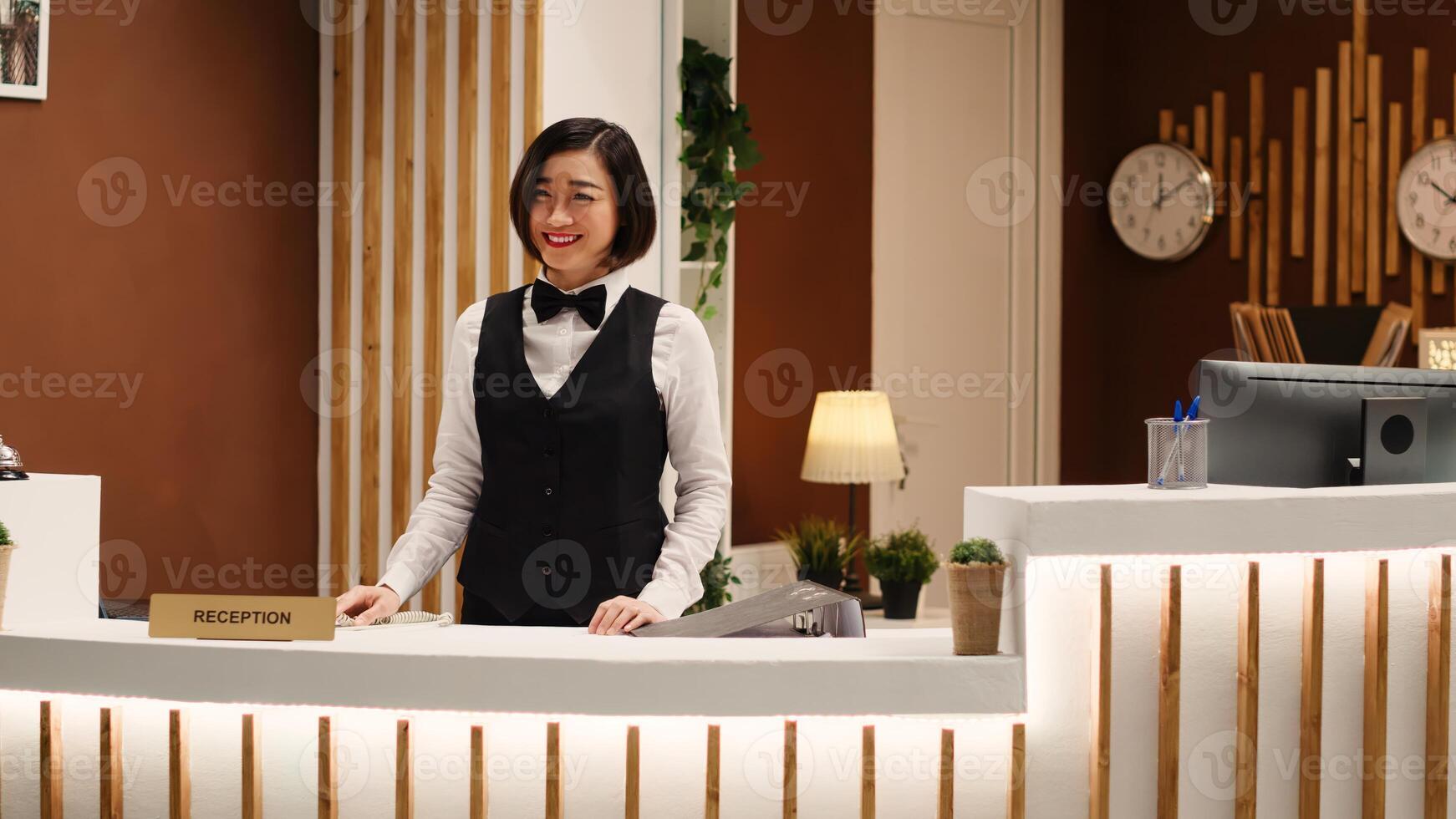 Smiling professional hotel employee taking phone calls while working at reception desk. Happy cheerful receptionist taking booking requests standing at check in counter in welcoming resort lobby photo