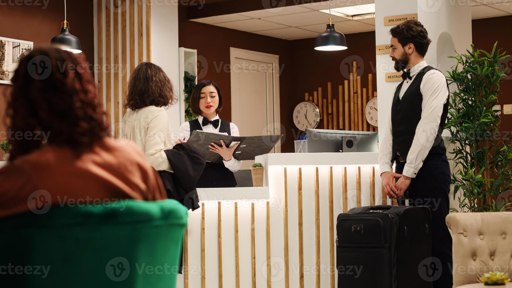 Tourist arriving in hotel lounge, assisted by helpful bellboy and receptionist managing her reservation details. Cheerful resort personnel maintaining positive attitude while helping customer photo