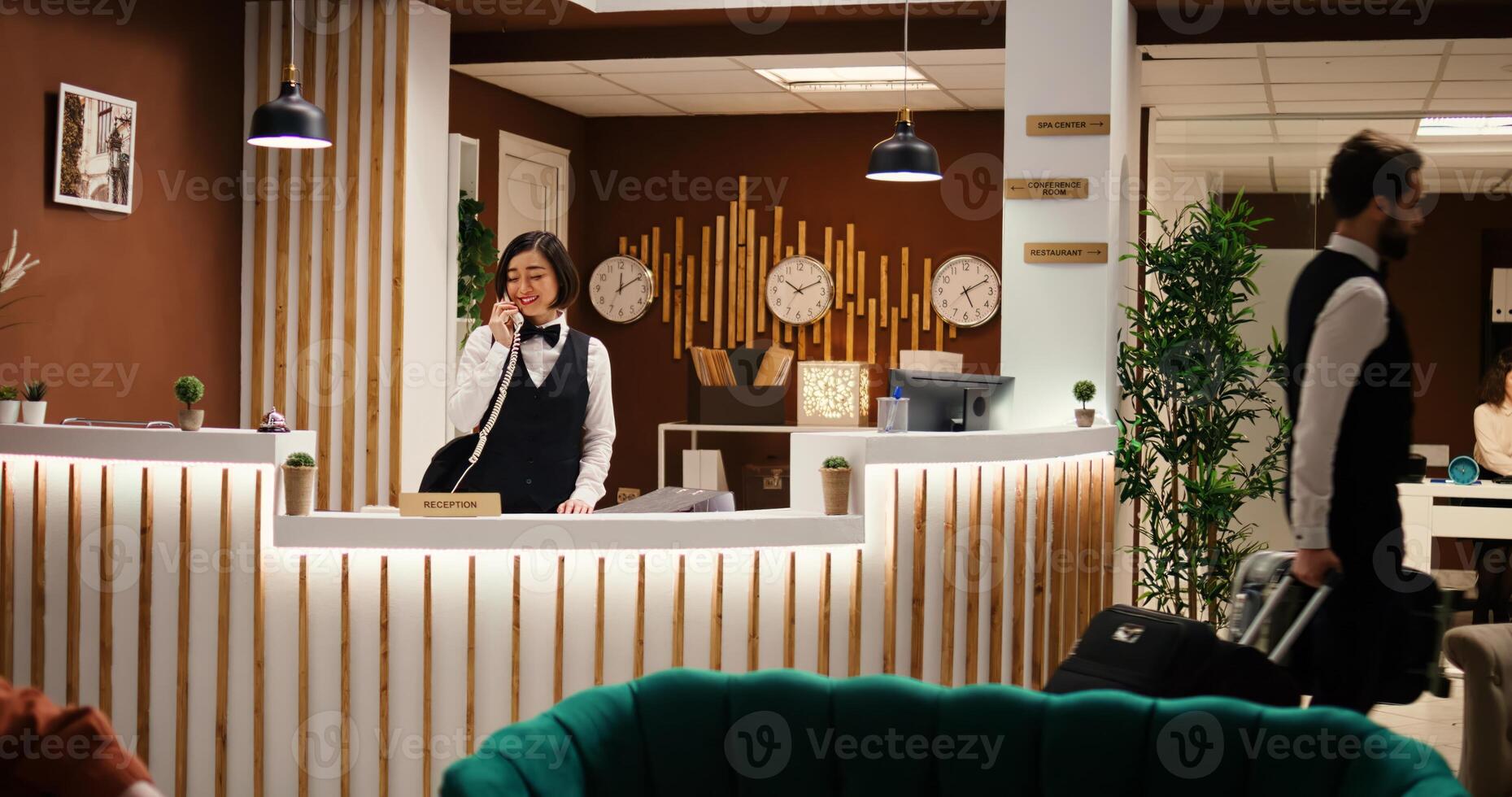 Happy professional hotel personnel doing various administrative tasks in stylish hotel reception. Asian receptionist taking room booking phone calls and bellboy carrying tourist luggage photo