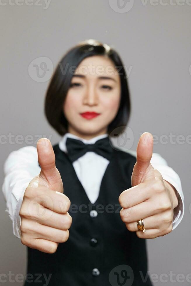 Cheerful asian woman in waitress uniform smiling and giving thumbs up symbol closeup. Cafe worker confirming and recommending restaurant good service and looking at camera photo