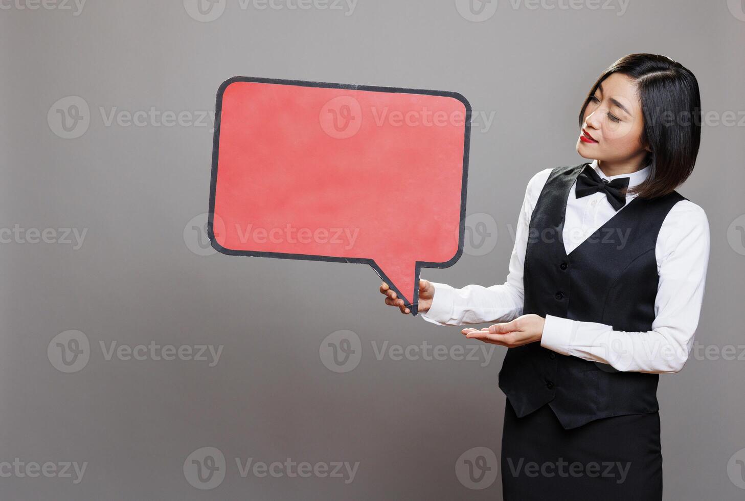 Young asian waitress holding blank chat balloon mockup while posing in studio. Attractive receptionist in professional uniform showing empty speak cloud with copy space for promo message photo