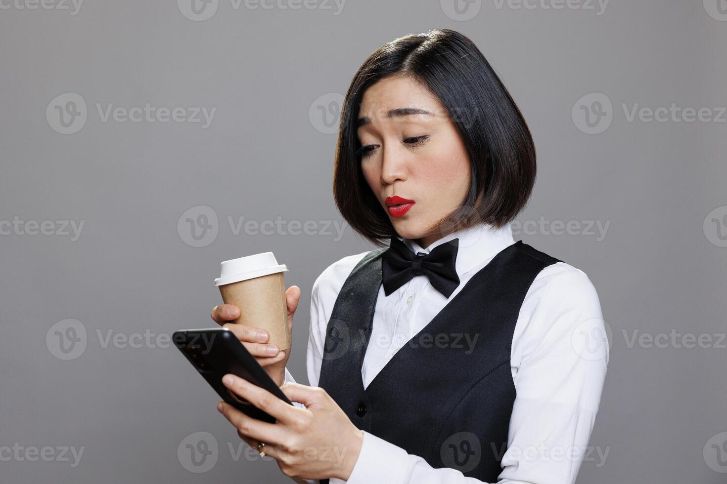 Asian woman in waitress uniform sending message on mobile phone during coffee break. Young receptionist talking on mobile phone social media and drinking hot beverage to go photo