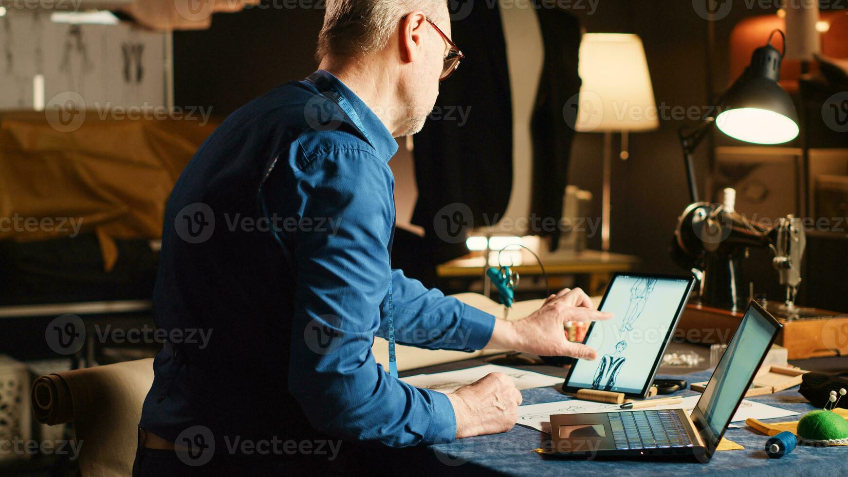 Old tailor works on clothes collection, comparing modern designs of tailored suit on laptop and tablet. Experienced fashion designer looking at sketches for new clothing line, craftsman job. photo