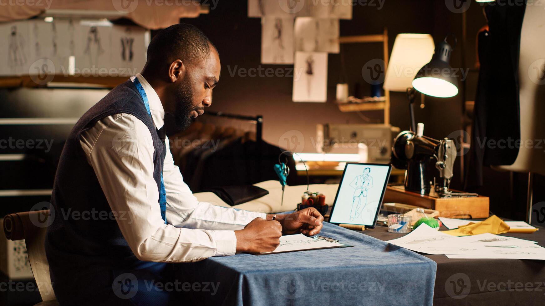 Couturier drawing sketches clothes at workstation, preparing to design new clothing line handmade. Fashion designer working on modern items, looking at models on tablet in atelier. photo