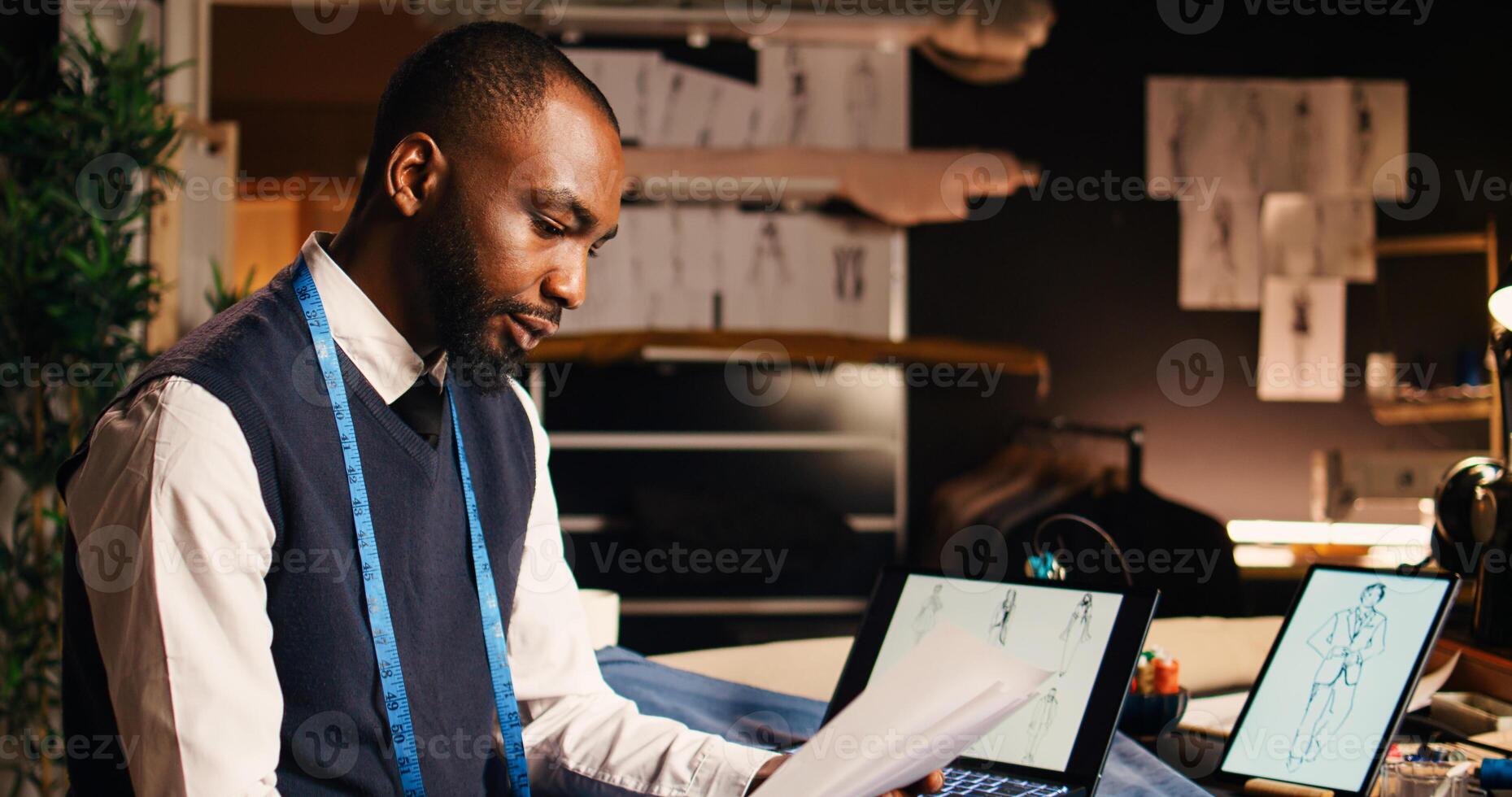 African american tailor checking sketches, using laptop and outlines on papers to compare fashion designs in atelier. Tailoring craftsman looking at clothing items models. Handheld shot. photo