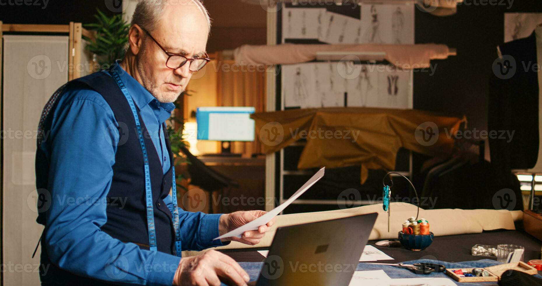 artesano mirando para diseño en computadora portátil, examinando bocetos con Moda artículos modelos experimentado Sastre diseño ropa en lujo taller, fabricación hecho a mano ropa línea. foto