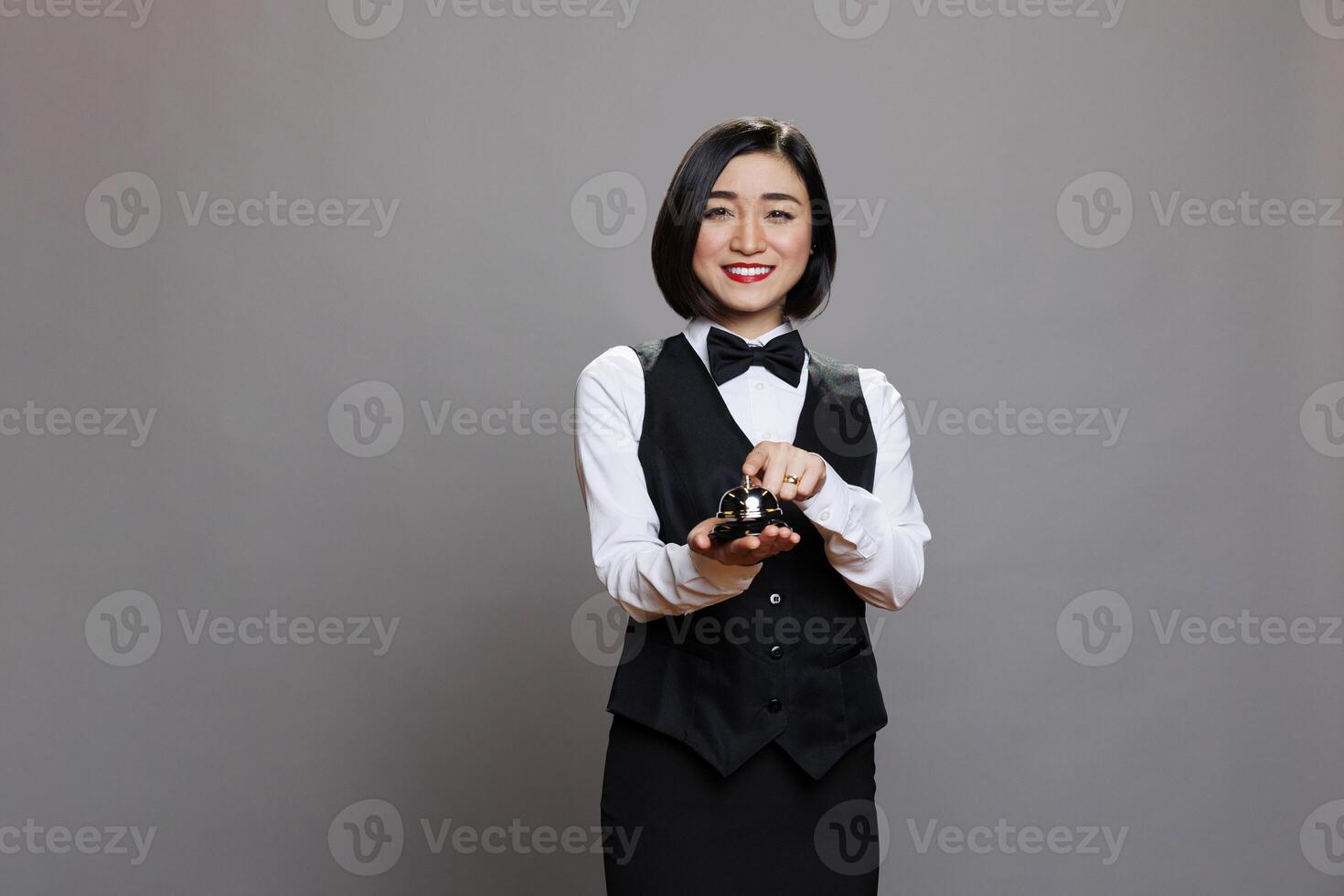 Smiling asian restaurant receptionist in uniform ringing dining bell while posing and looking at camera. Young waitress pressing order notification alert portrait on gray background photo