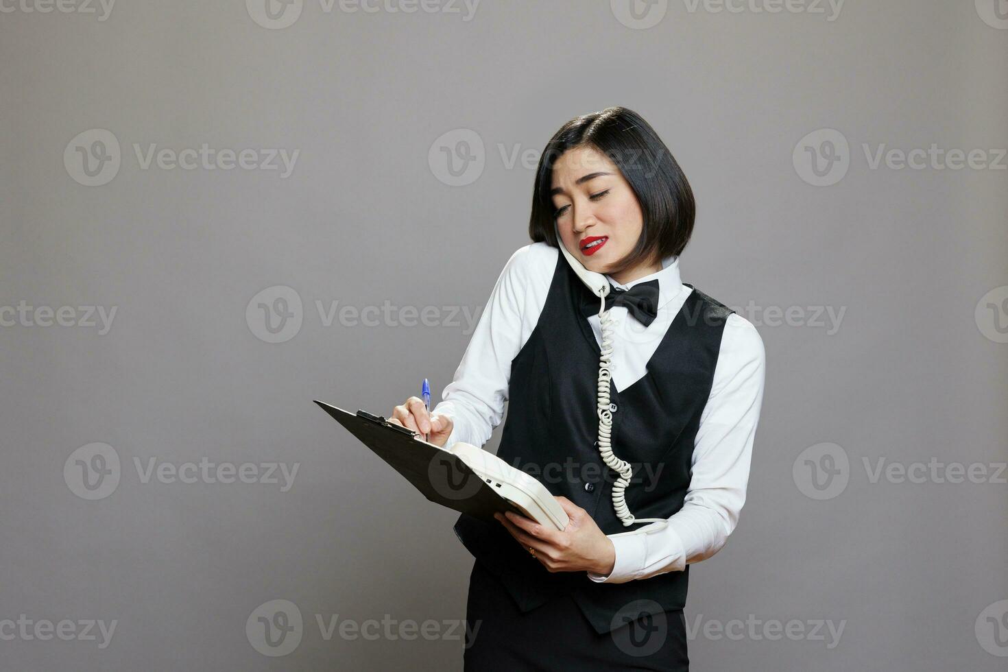 Waitress holding clipboard and talking on landline phone, managing communication and tasks in cafe. Receptionist engaging in conversation on telephone and writing information photo