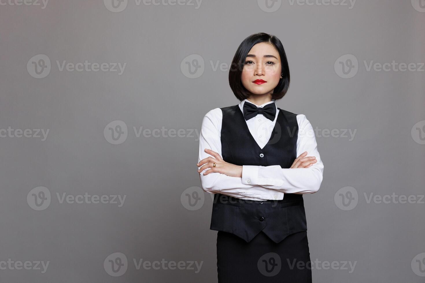 Catering service young asian waitress posing with crossed arms showcasing professionalism and looking at camera. Restaurant receptionist wearing black and white uniform portrait photo