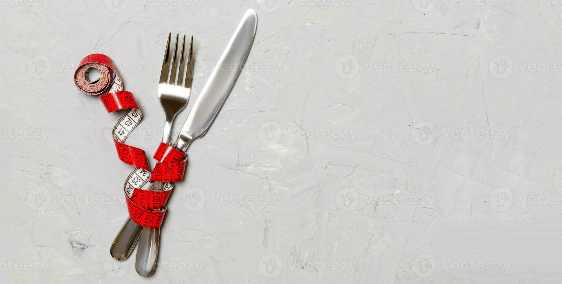 Crossed fork and knife are wrapped in measuring tape on gray background. Concept of diet for weight loss with copy space photo