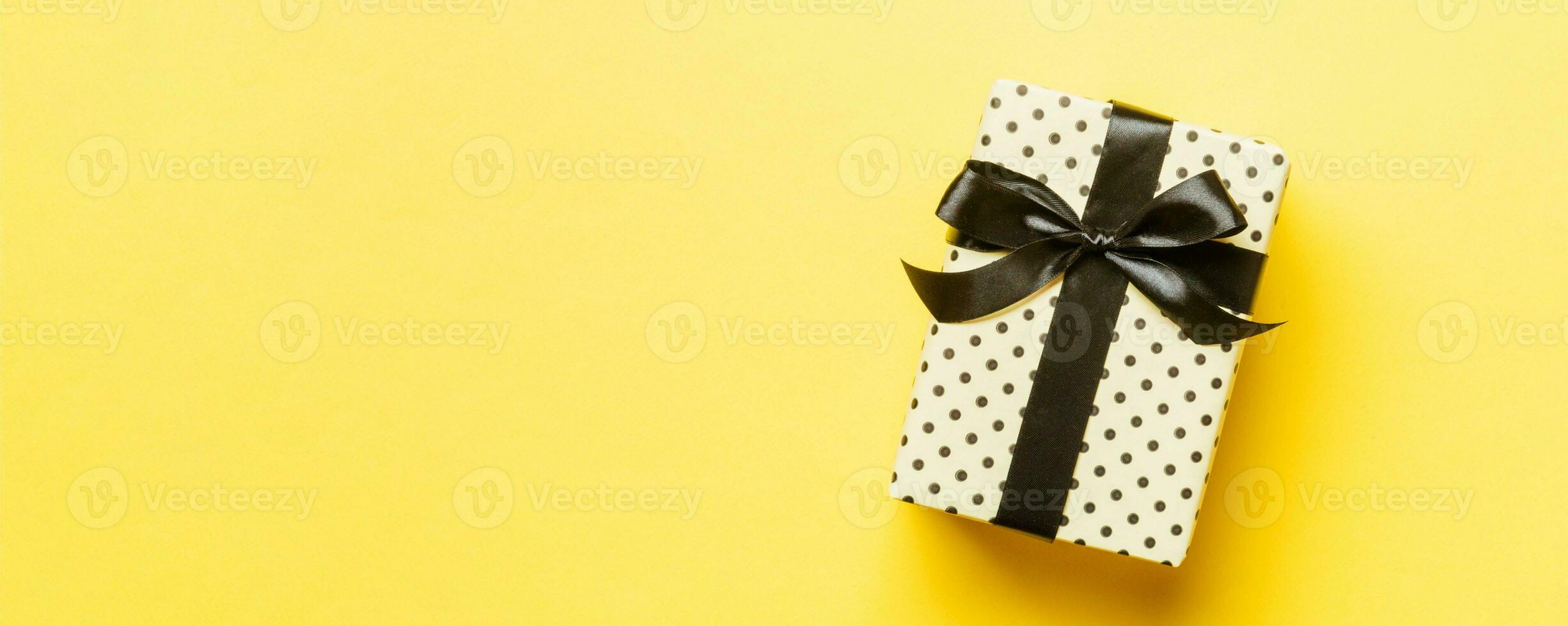 wrapped Christmas or other holiday handmade present in paper with black ribbon on yellow background. Present box, decoration of gift on colored table, top view with copy space photo