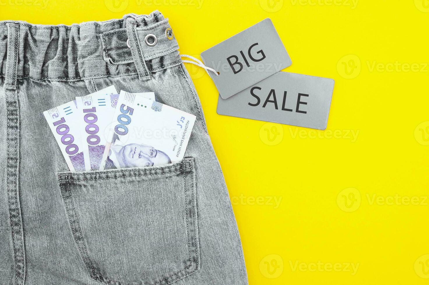 Flat lay, detail of denim jeans with Ukrainian hryvnia bills in pocket with gray label and inscription SALE. photo