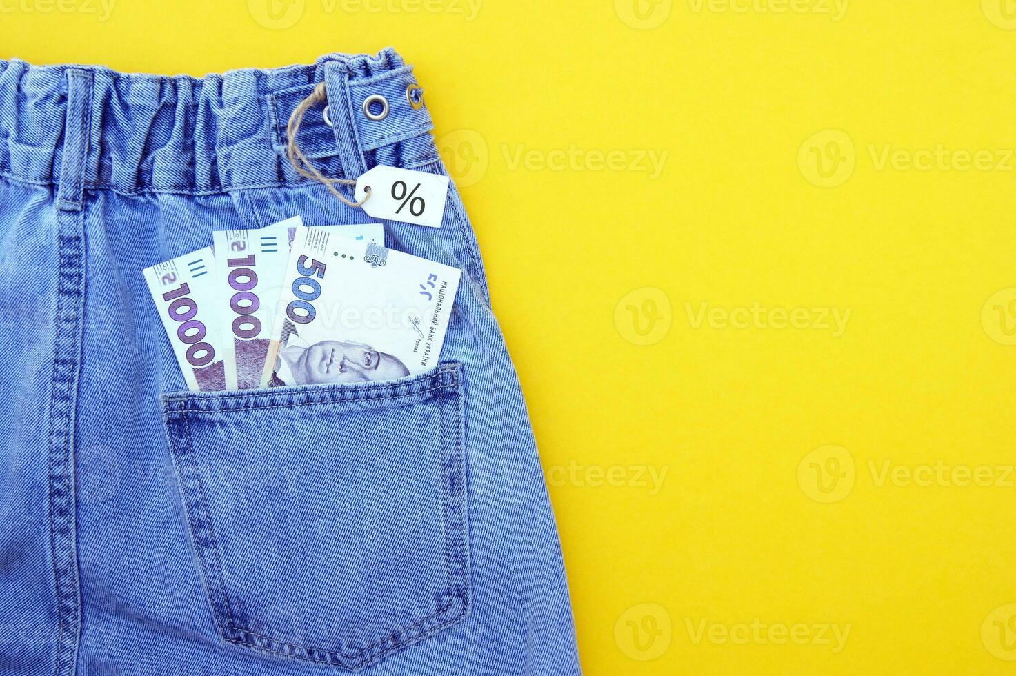 Flat lay, detail of denim jeans with Ukrainian hryvnia bills in pocket with white label on yellow background. photo