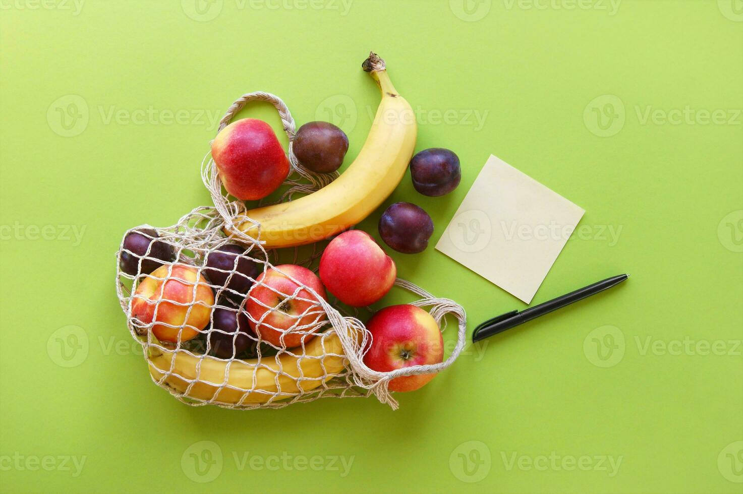 Red apples, bananas, plums, string bag and blank paper with black pen on a bright green background. photo