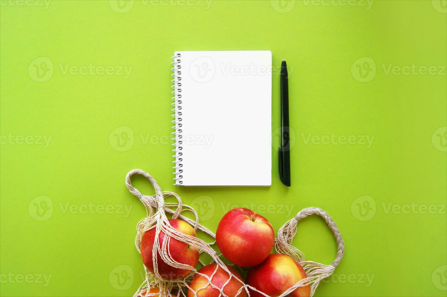 Red apple in string bag and notebook with pen on a bright green background. photo