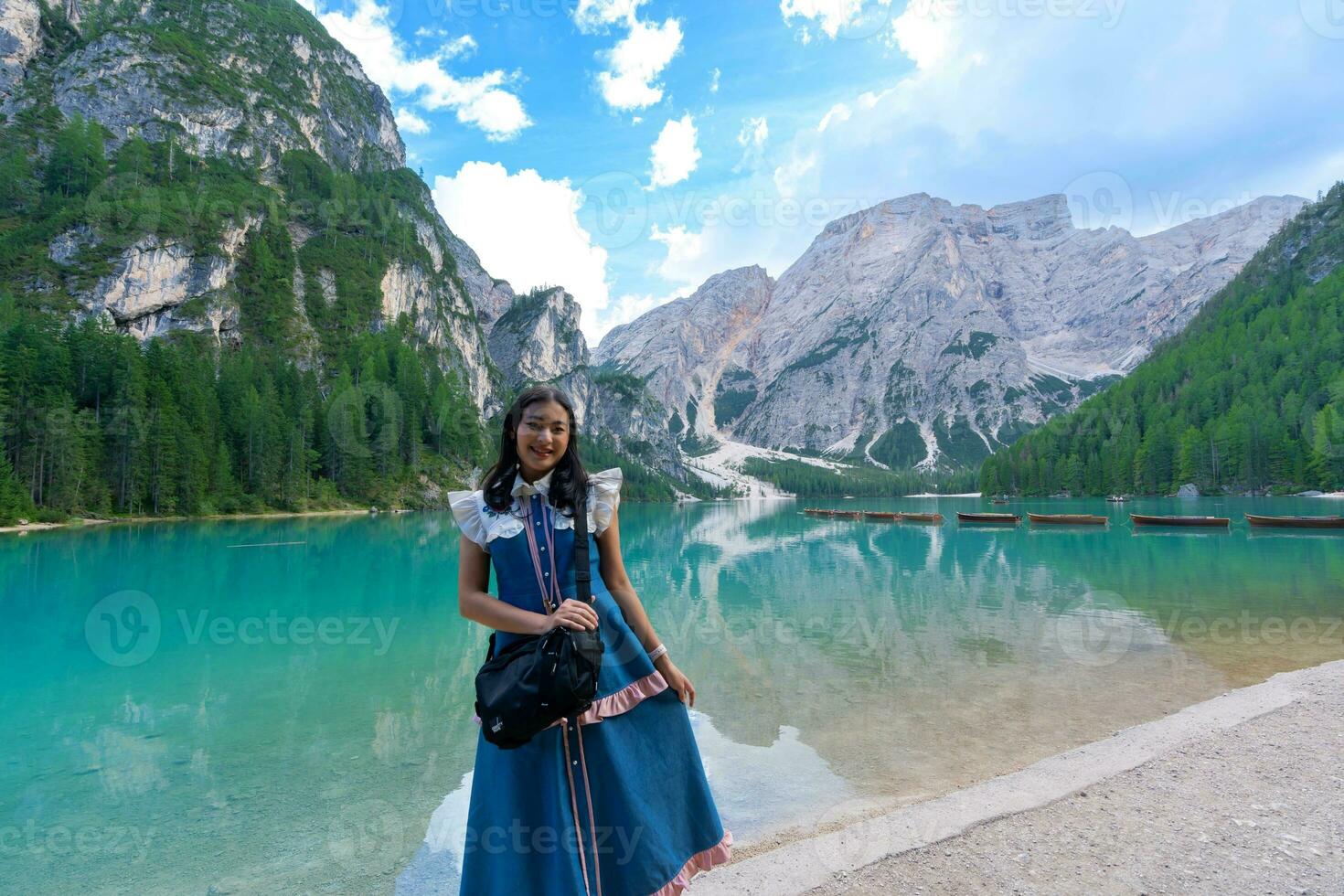 happy Asian woman tourist standing on the coast of Braies lake. Dolomites, Italy. Landscape with girl, famous lake with beautiful reflection in water, trees, sky with clouds. Travel. photo