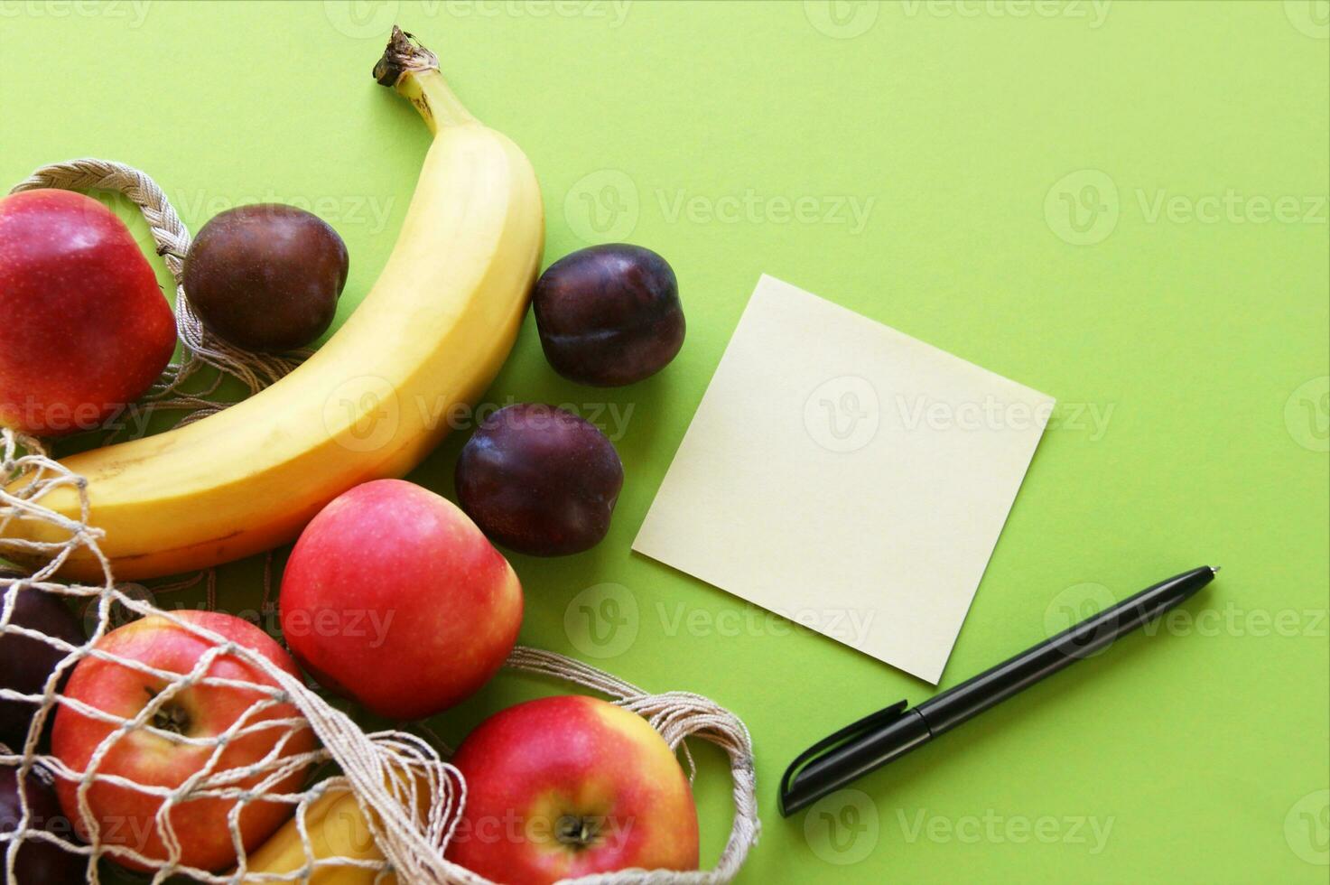 Red apples, bananas, plums, string bag and blank paper with black pen on a bright green background. photo