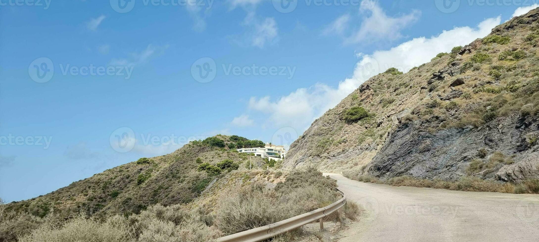 Winding road leading to a hillside house with clear blue skies and sparse vegetation. photo
