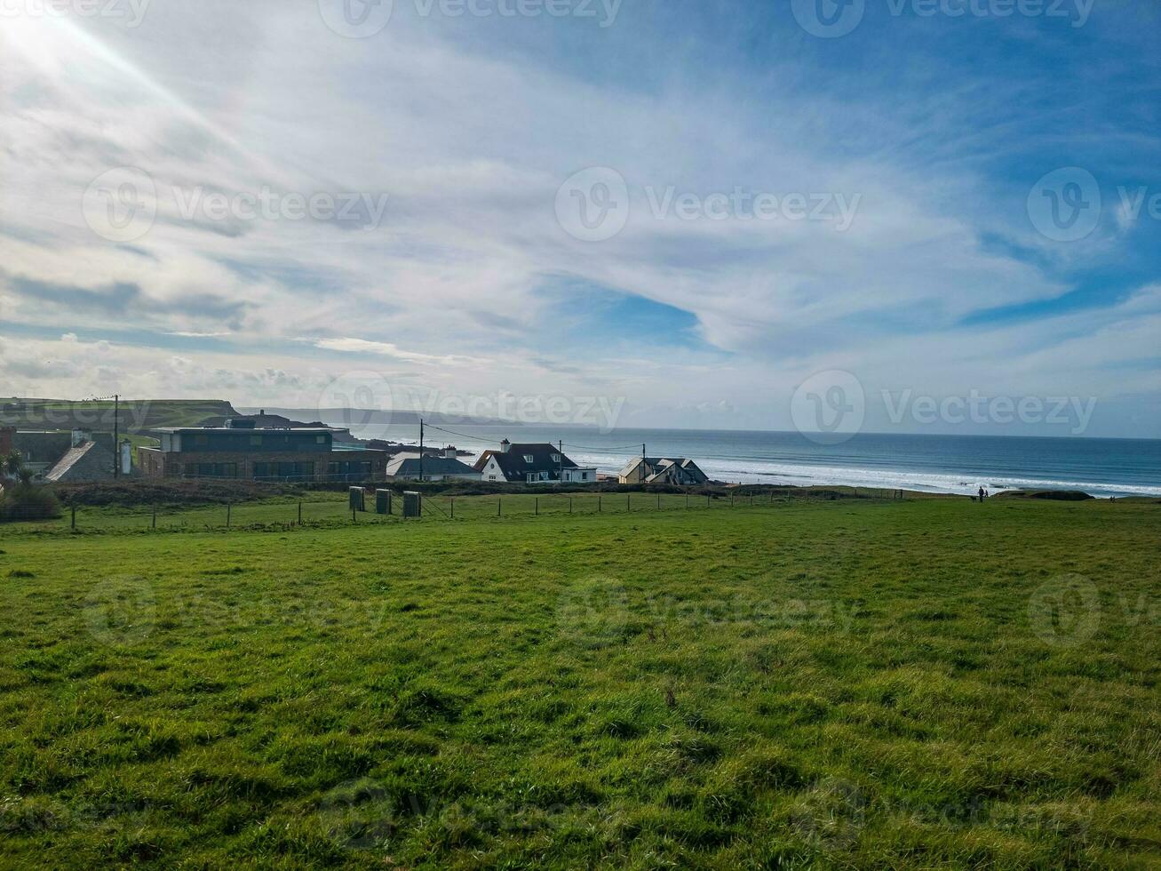 Coastal landscape with green fields, houses, and a distant beach under a blue sky with clouds. photo