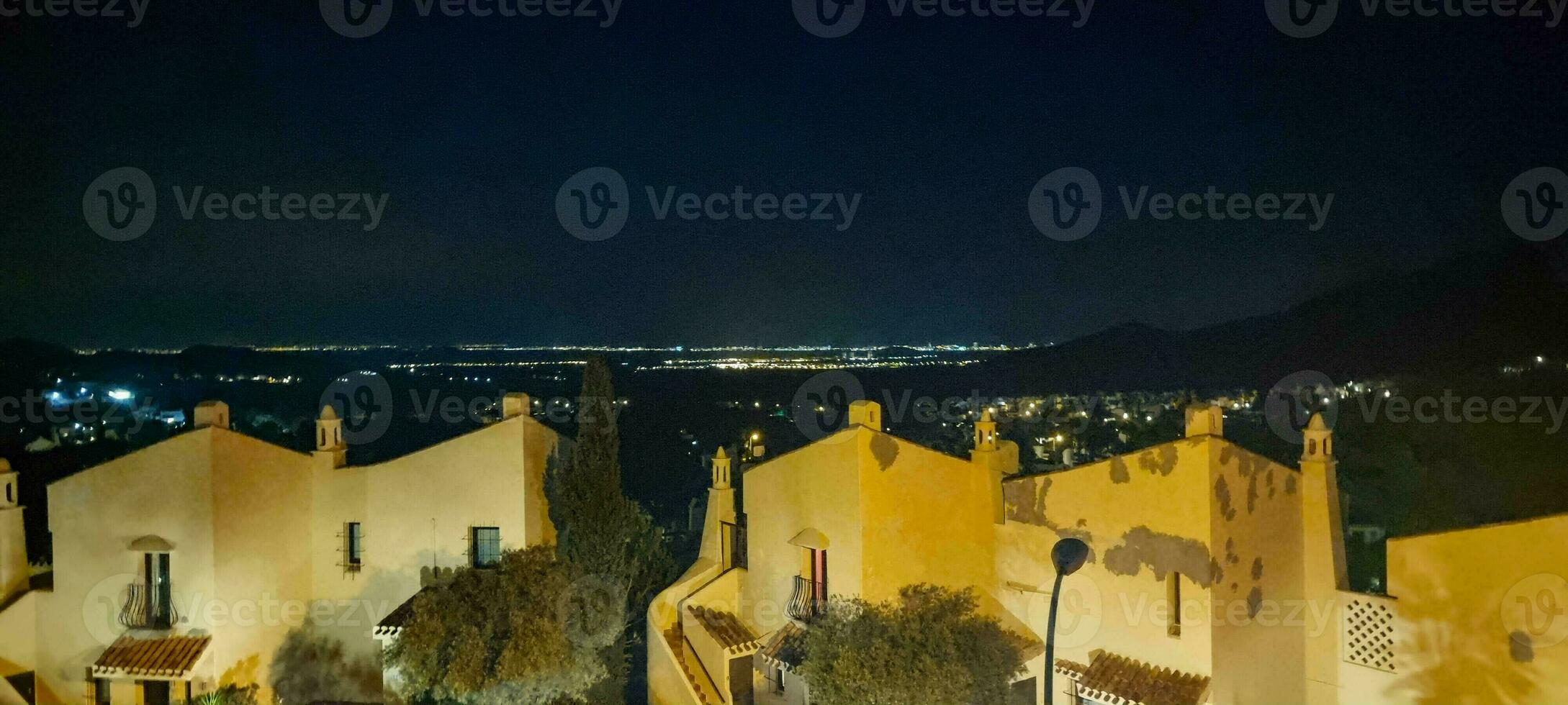 Night view of traditional buildings with illuminated windows against a backdrop of distant city lights and mountains. photo