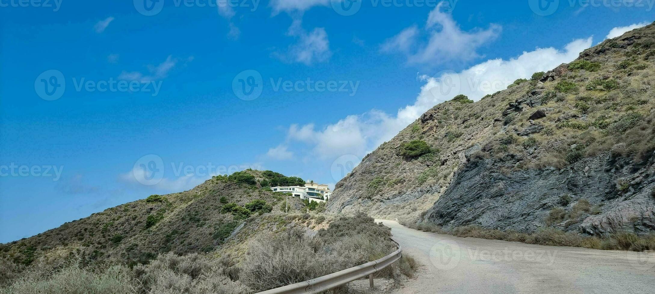 Winding mountain road with clear blue sky and sparse vegetation leading to a distant white building. photo