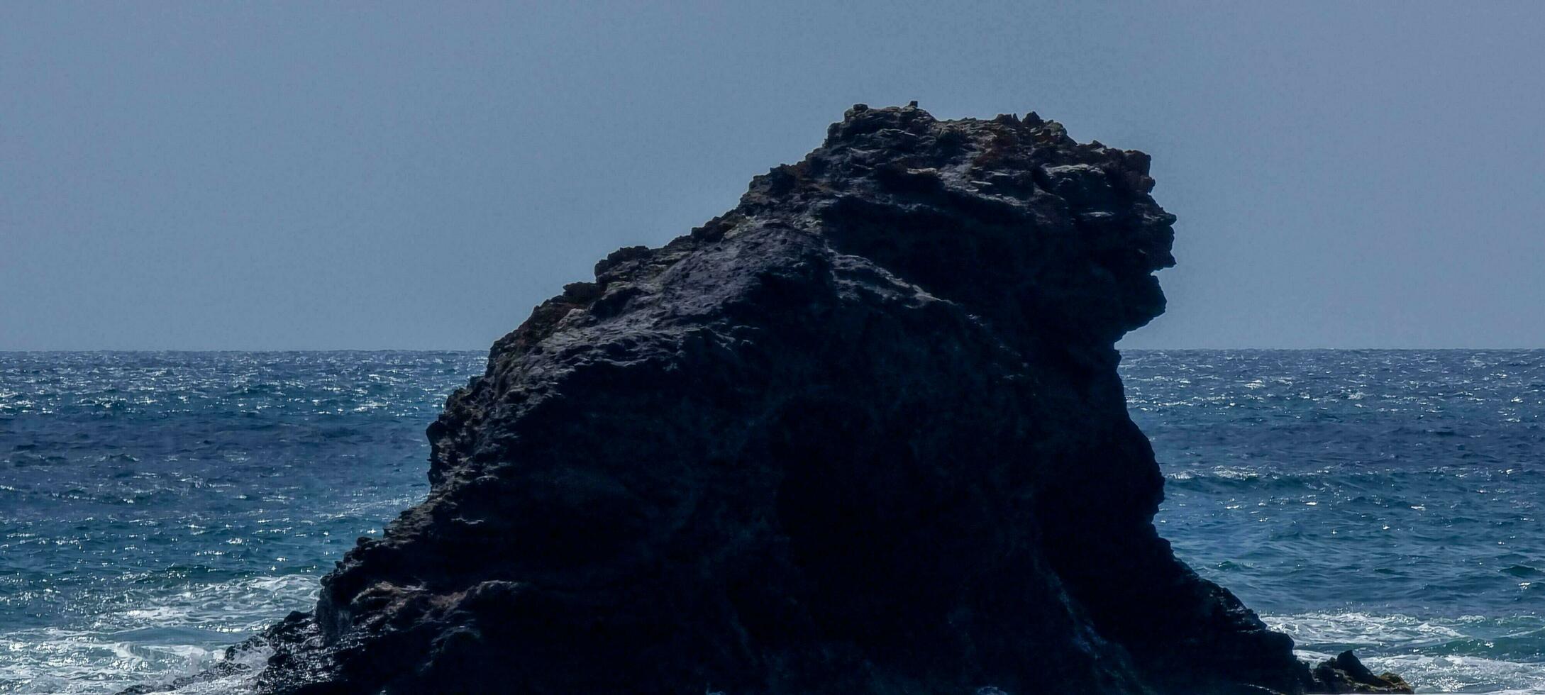 Large volcanic rock formation against a calm blue ocean under a clear sky. photo