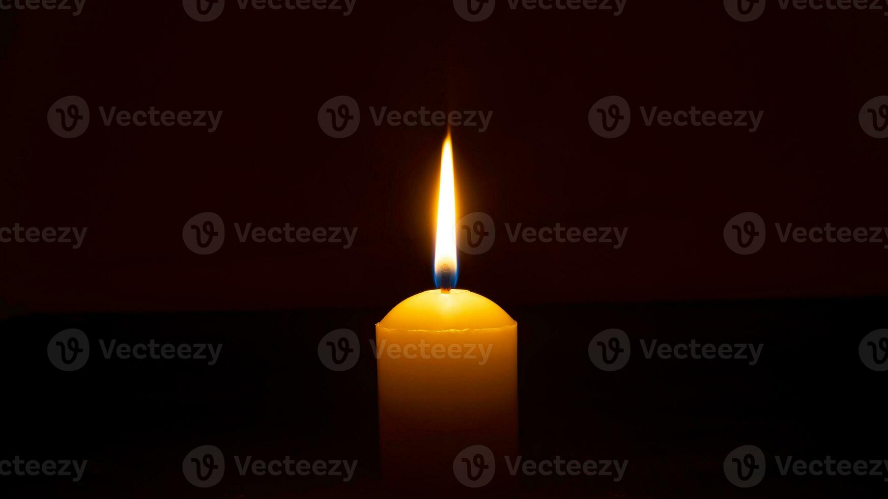 A single burning candle flame or light glowing on a big yellow candle isolated on red or dark background on table in church for Christmas, funeral or memorial service photo