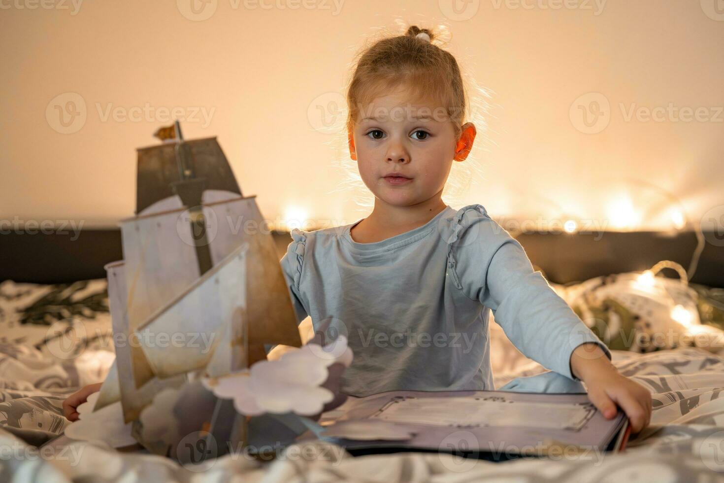 Little girl reading pop up book in home bed in Christmas environment with lights background. High quality photo