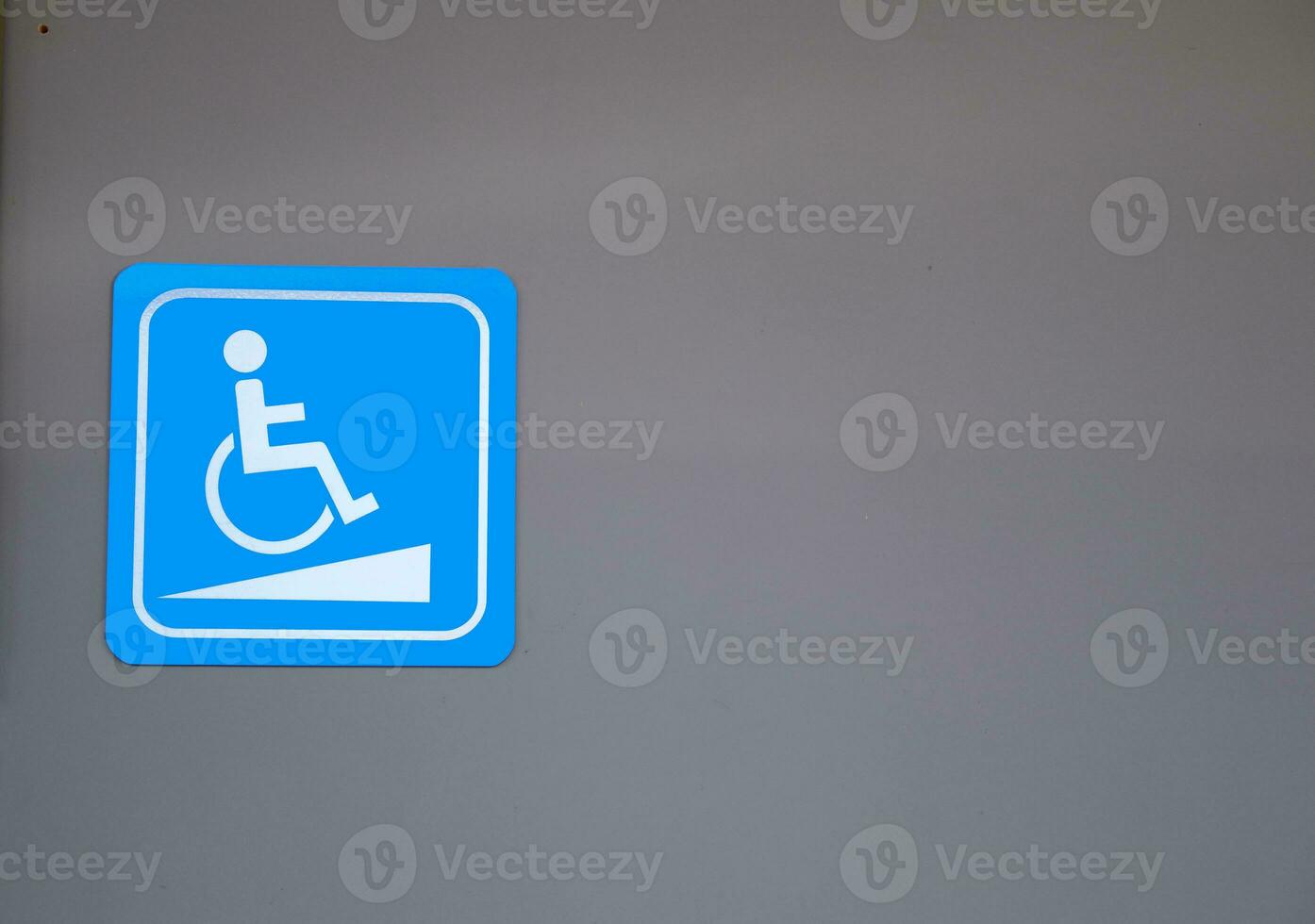 White sign of handicapped ramp for wheelchair on bright blue square shape on gray wall of building. photo