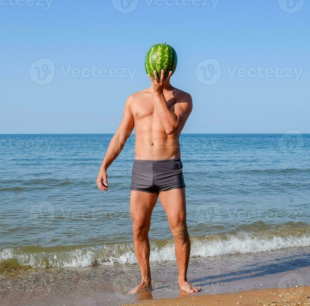 A sporty man is standing on the beach and holding a whole watermelon. A ripe watermelon in the hands of a man photo