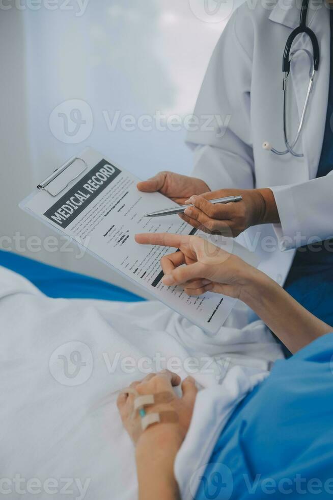 The senior woman specialist doctor with stethoscope checking up happy young Asian female patient lying in bed with receiving an intravenous saline drip in recovery room. Healthcare, medical insurance. photo