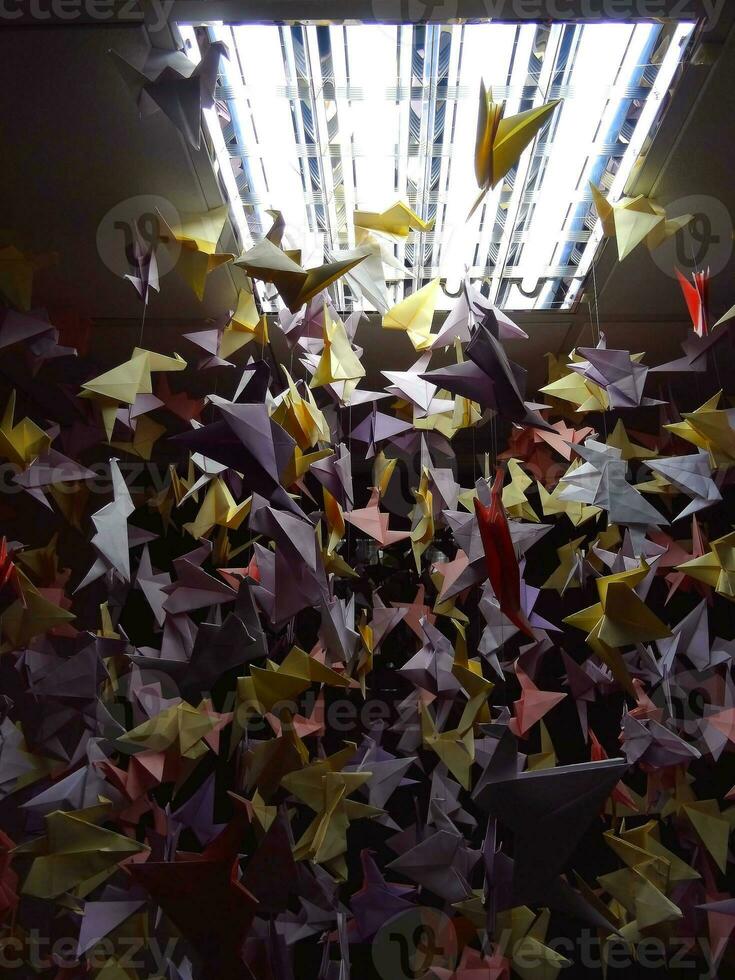 Handmade colored origami cranes on ceiling strings under light lamp photo