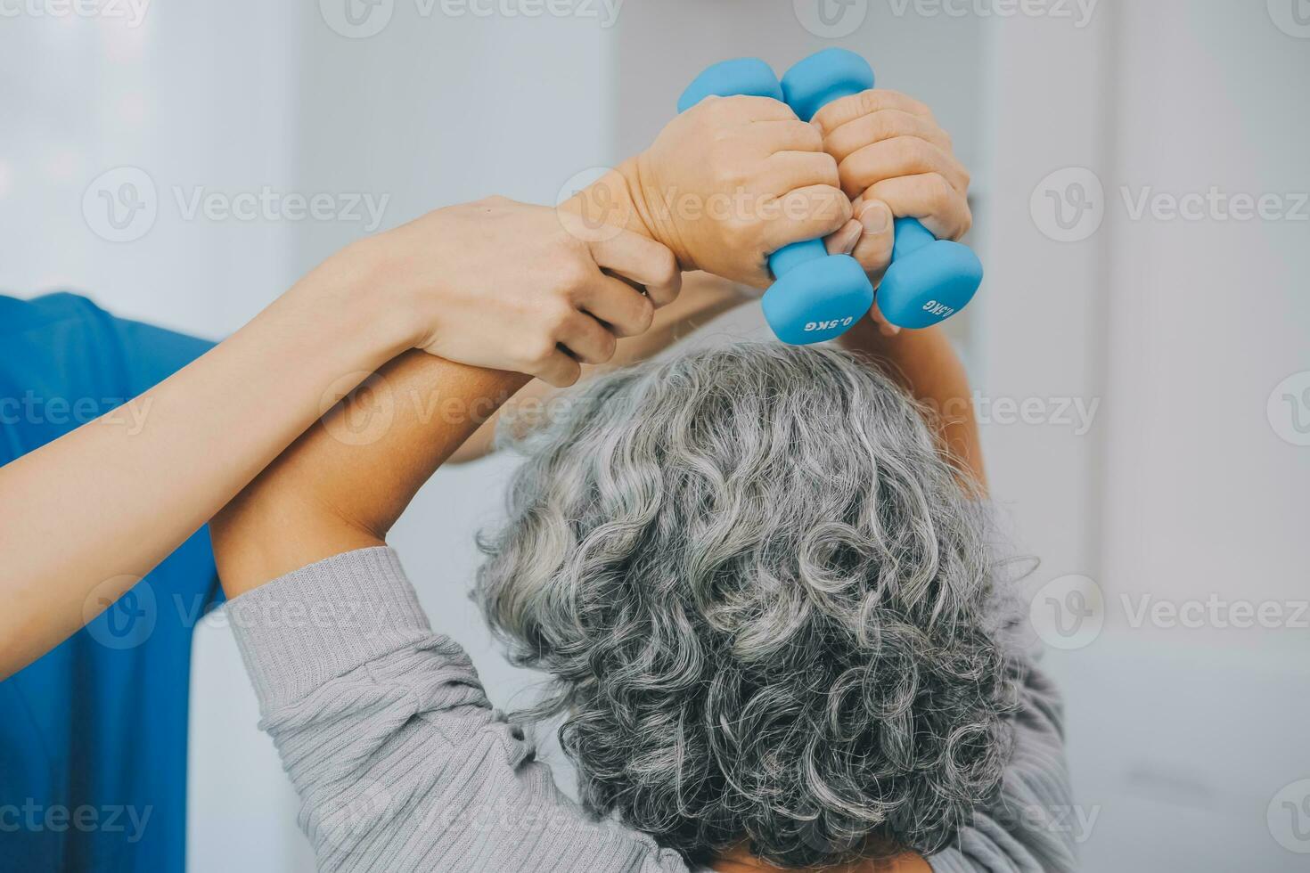 Old woman training with physiotherapist using dumbbells at home. Therapist assisting senior woman with exercises in nursing home. Elderly patient using dumbbells with outstretched arms. photo