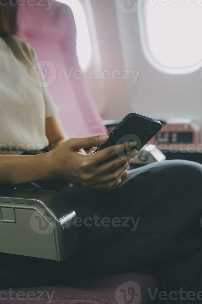 Asian woman passenger sitting in airplane near window and reading news from social networks or using travel applications in smartphone photo