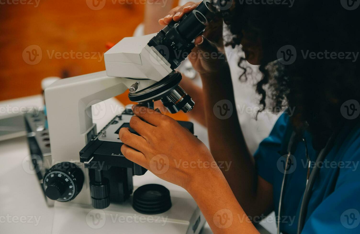 Medical worker in lab coat and sterile mask, doing a microscope analysis while her colleague are working behind photo