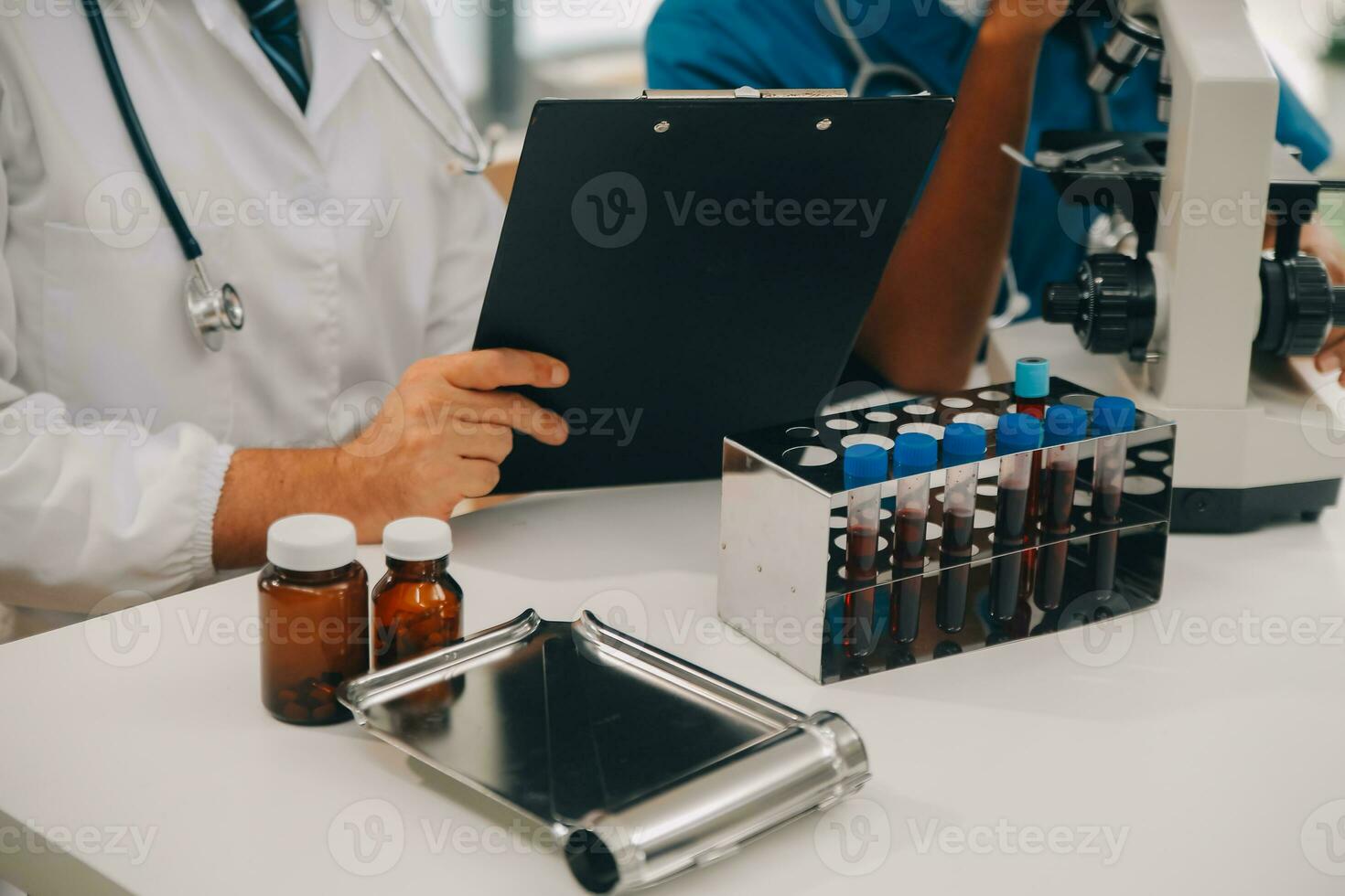 Medical worker in lab coat and sterile mask, doing a microscope analysis while her colleague are working behind photo