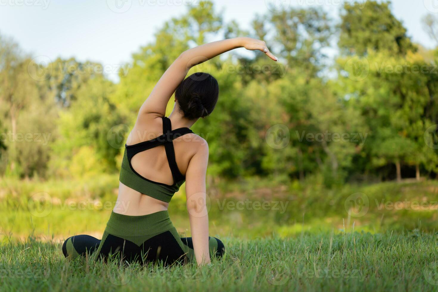 View from the back. Sitting on the grass, a teenager girl performs tilts to the sides with a beautiful view of the summer nature. Physical form, harmony photo